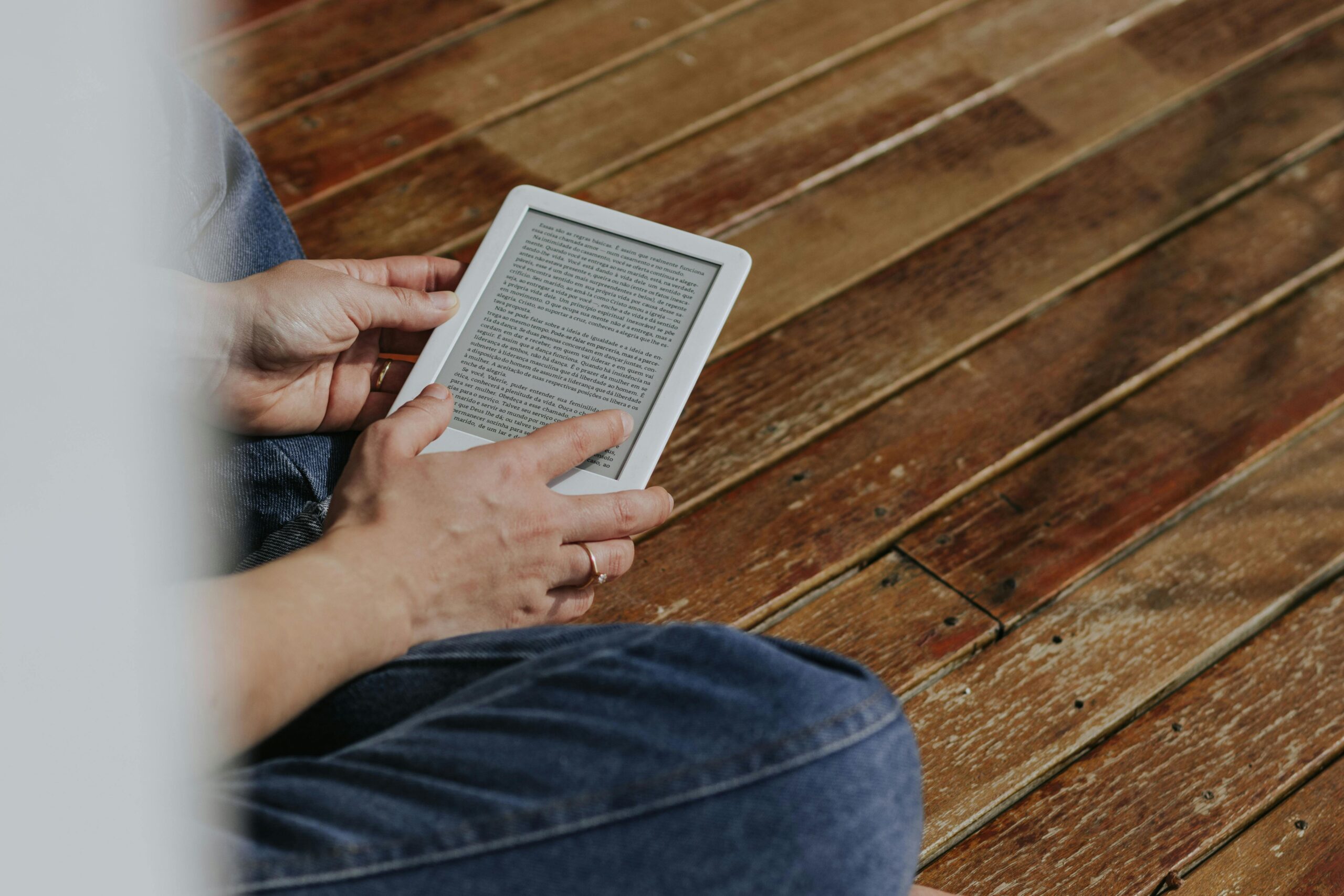 Close-up of hands holding an e-reader on a wooden floor, depicting a casual reading moment.
